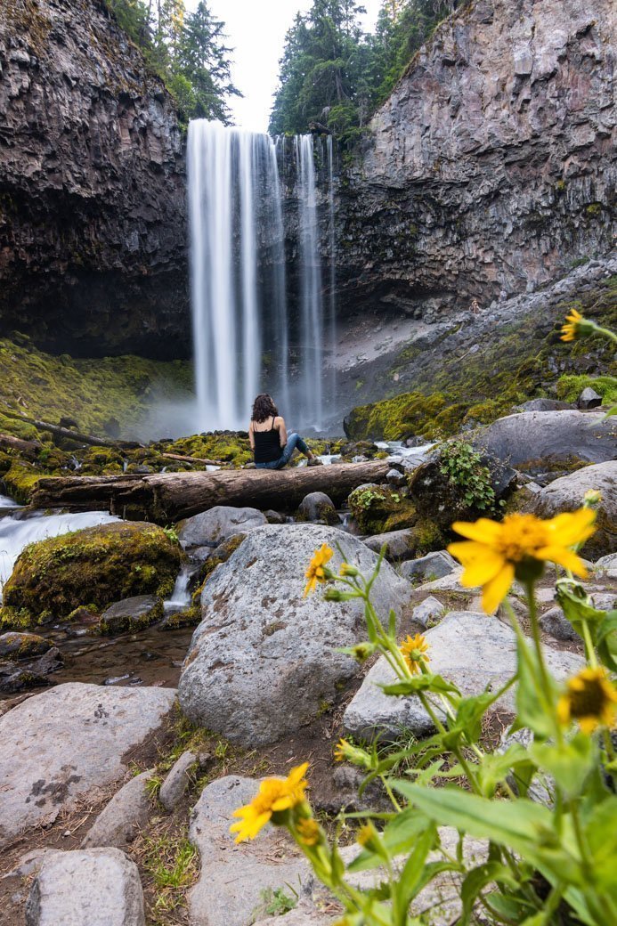 Tamanawas Falls tumbling over a cliff with Nina sitting in front on a piece of driftwood.