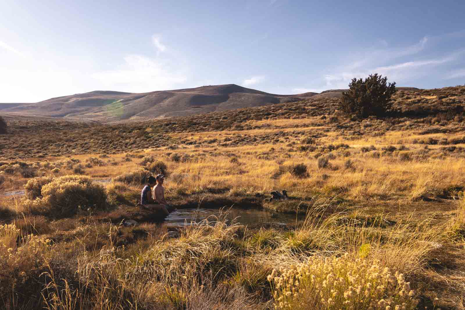 Two people bathing inside the natural Hart Mountain Hot Spring with views across the meadow and hills.