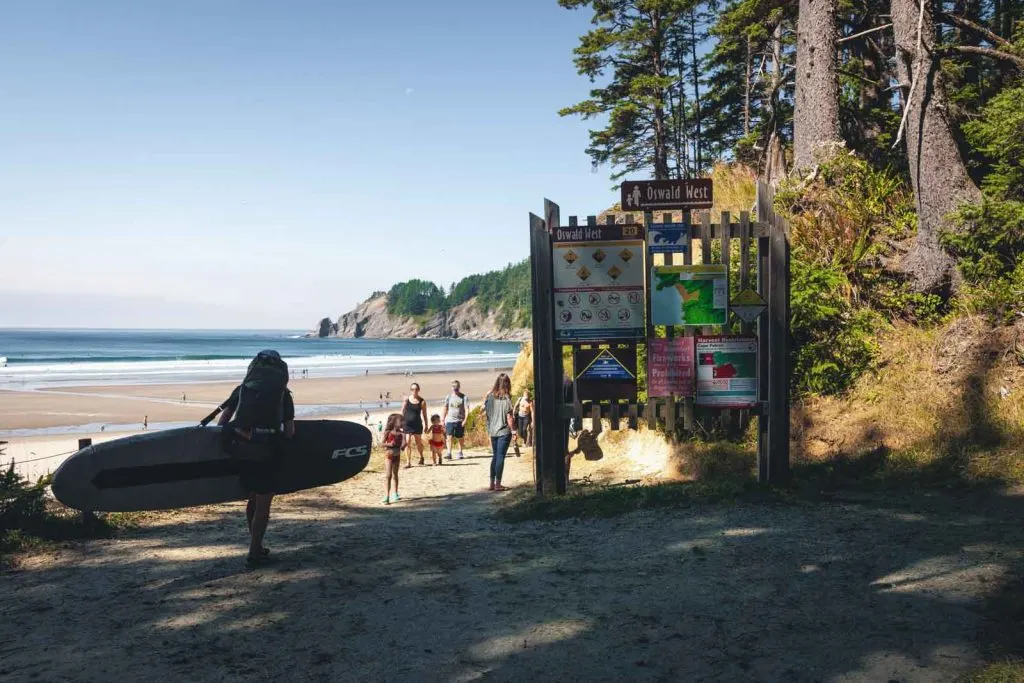 Person with surfboard by large sign with beach in the background at Oswald West, one of the Oregon Coast State Parks
