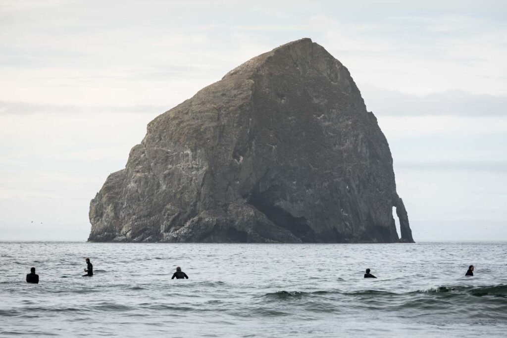 Surfers in ocean with large rock island in background at Cape Kiwanda, one of the best beaches on the Oregon Coast