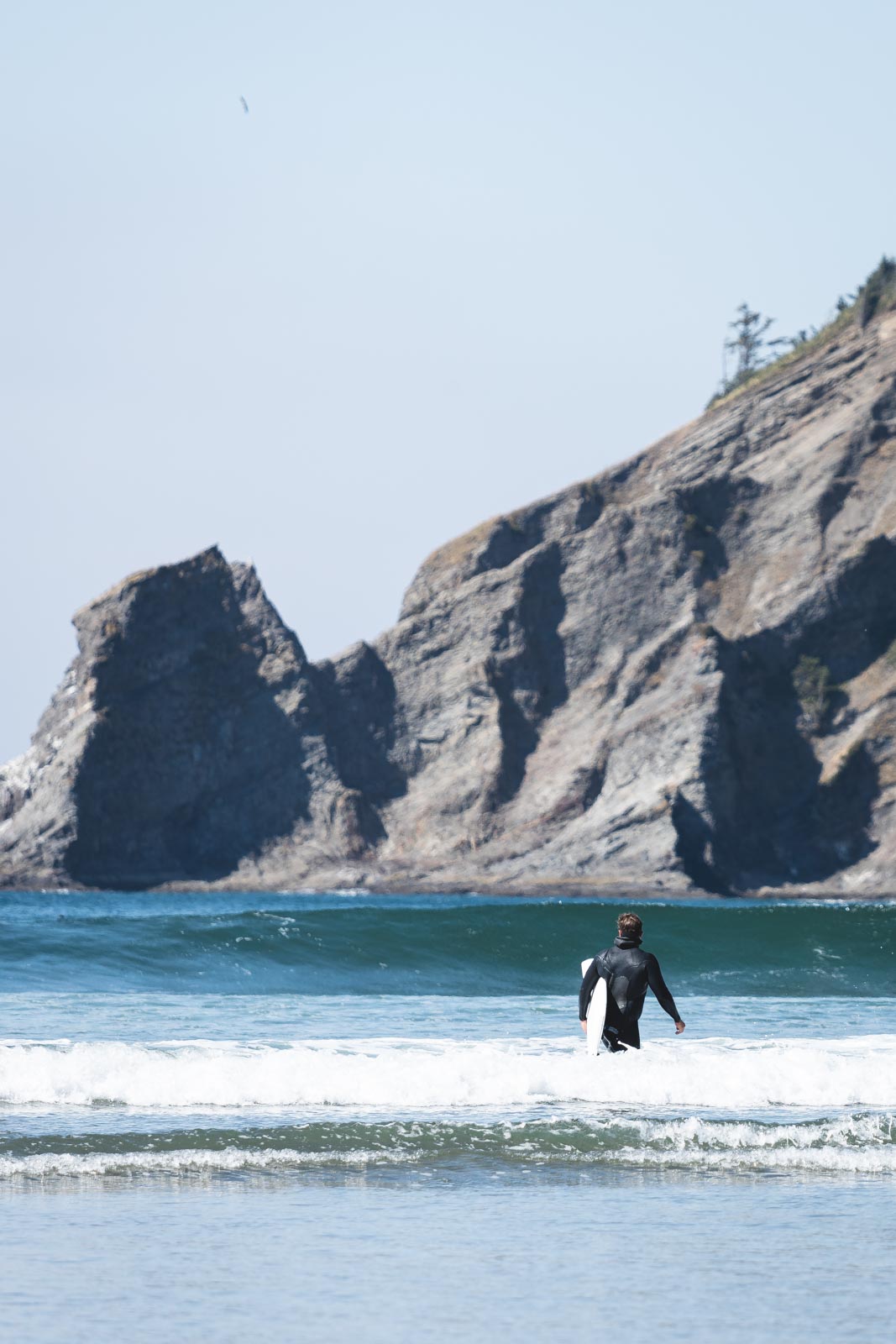 Man holding surfboard in ocean with rocky cliffs in background at Oswald West State Park.