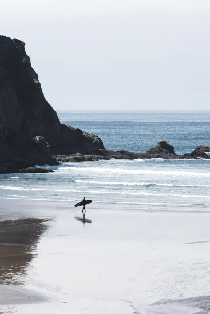 Man walking with surfboard on the beach with rocky cliffs in background at Oswald West State Park