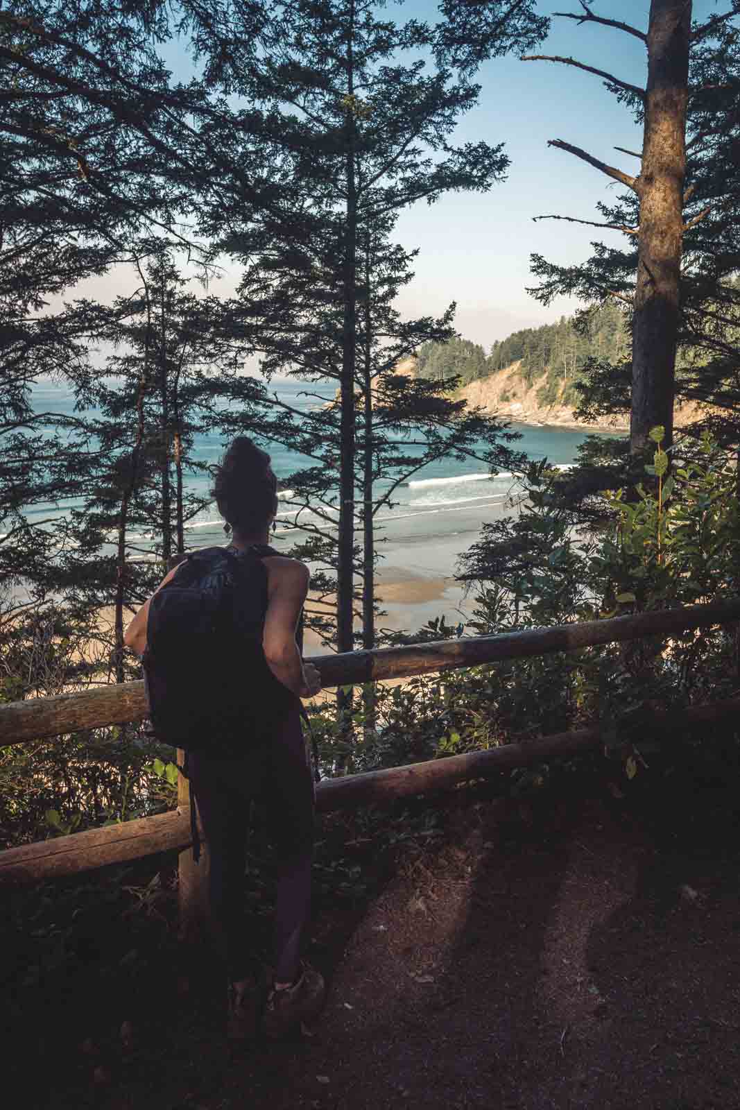 Woman hiking on trail surrounded by trees and with view through trees of coast in Oswald West State Park