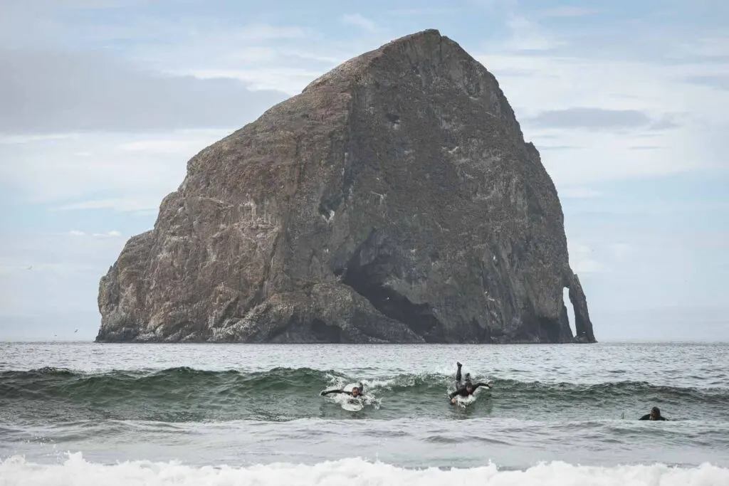 Two surfers surfing wave in ocean in front of large rock island in Pacific City, Oregon