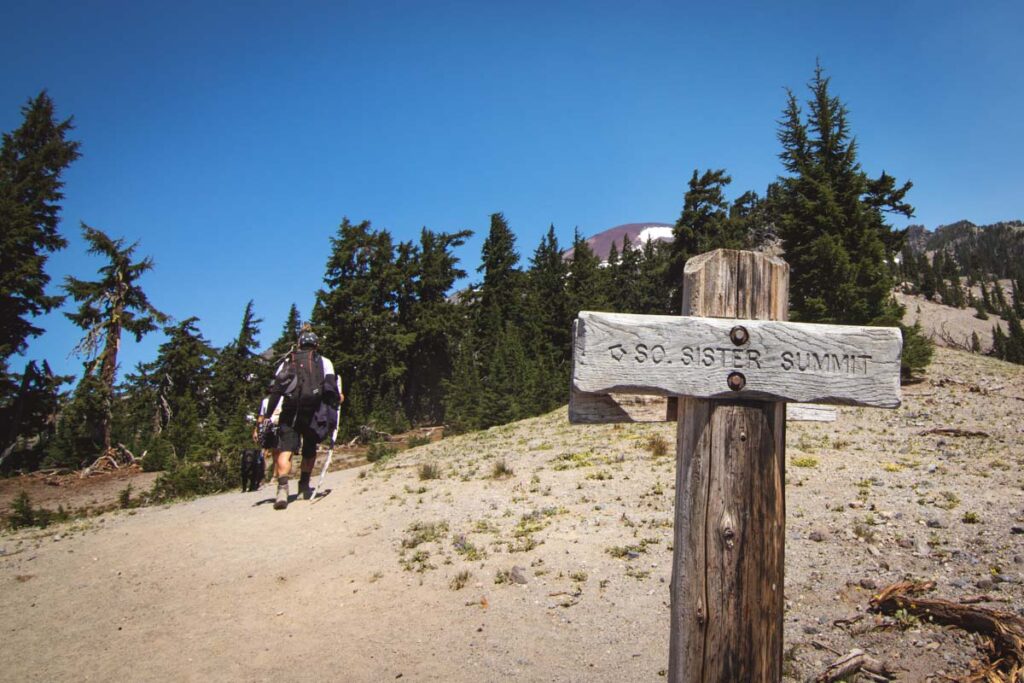 Trail sign for hiking south sister trail with two people hiking on trail into trees