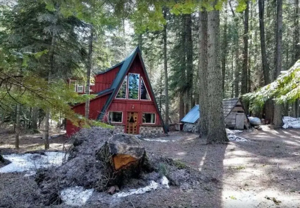 Red A-Frame cabin in Oregon surrounded by trees with large tree stump in foreground 