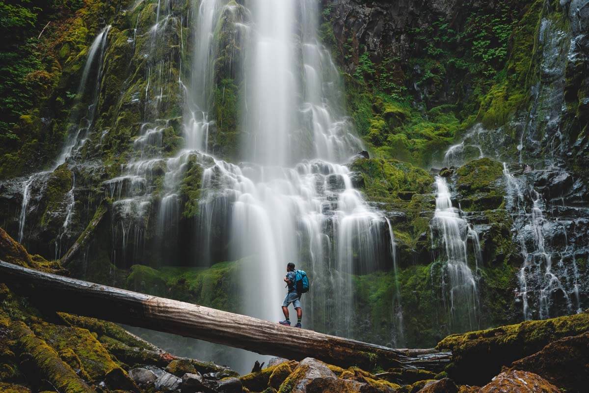 Garrett standing on fallen tree in front of Proxy Falls waterfall in the Oregon forest.