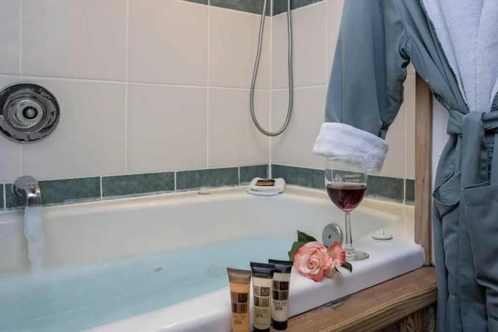 A glass of red wine, a pink rose and bathrobe next to a full bathtub in the bathroom of the Clementine's Guest House in Astoria.