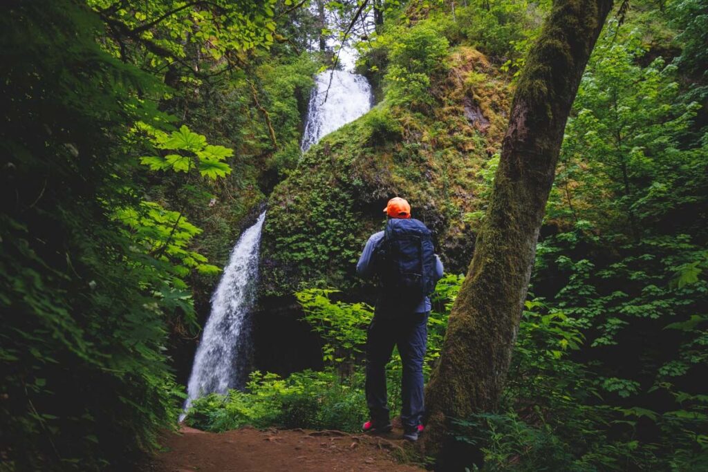 A male hiker standing beneath Latourell Falls surrounded by green forest foliage in Columbia River Gorge.