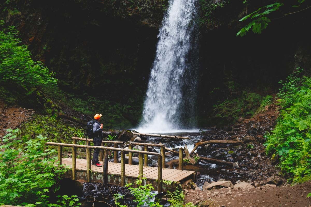 A male hiker standing on a wooden bridge in front of the base of Latourell Falls in Oregon.