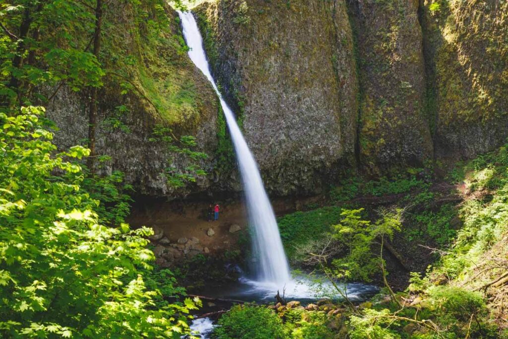 Two hikers stopped on a trail embedded in the cliffside behind Ponytail Falls.