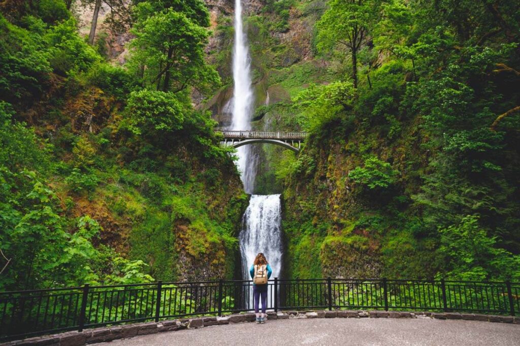 Female tourists stands at the base of Multnomah Waterfalls looking up at the cascade and bridge surrounded by trees.