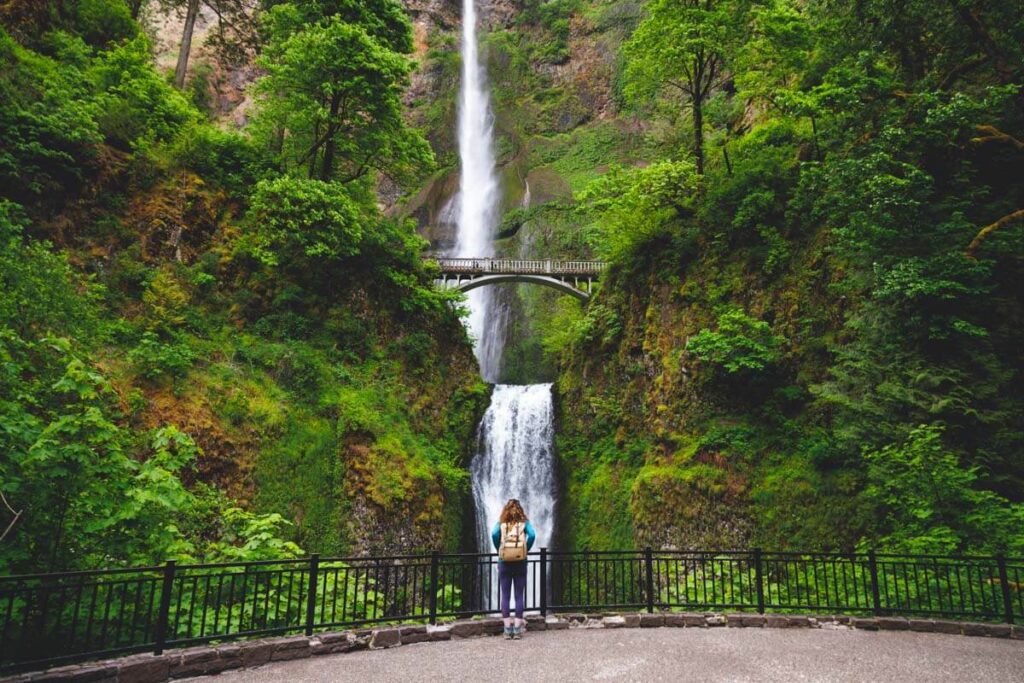 Female tourists stands at the base of Multnomah Waterfall looking up at the cascade and bridge surrounded by trees.