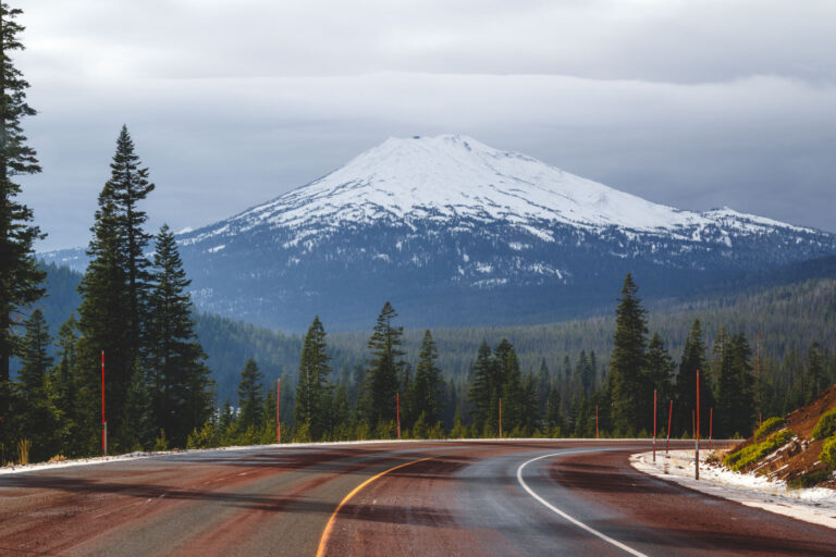 A reddish road curving past a snow-capped Mount Bachelor which is surrounded by a forest on a cloudy day.