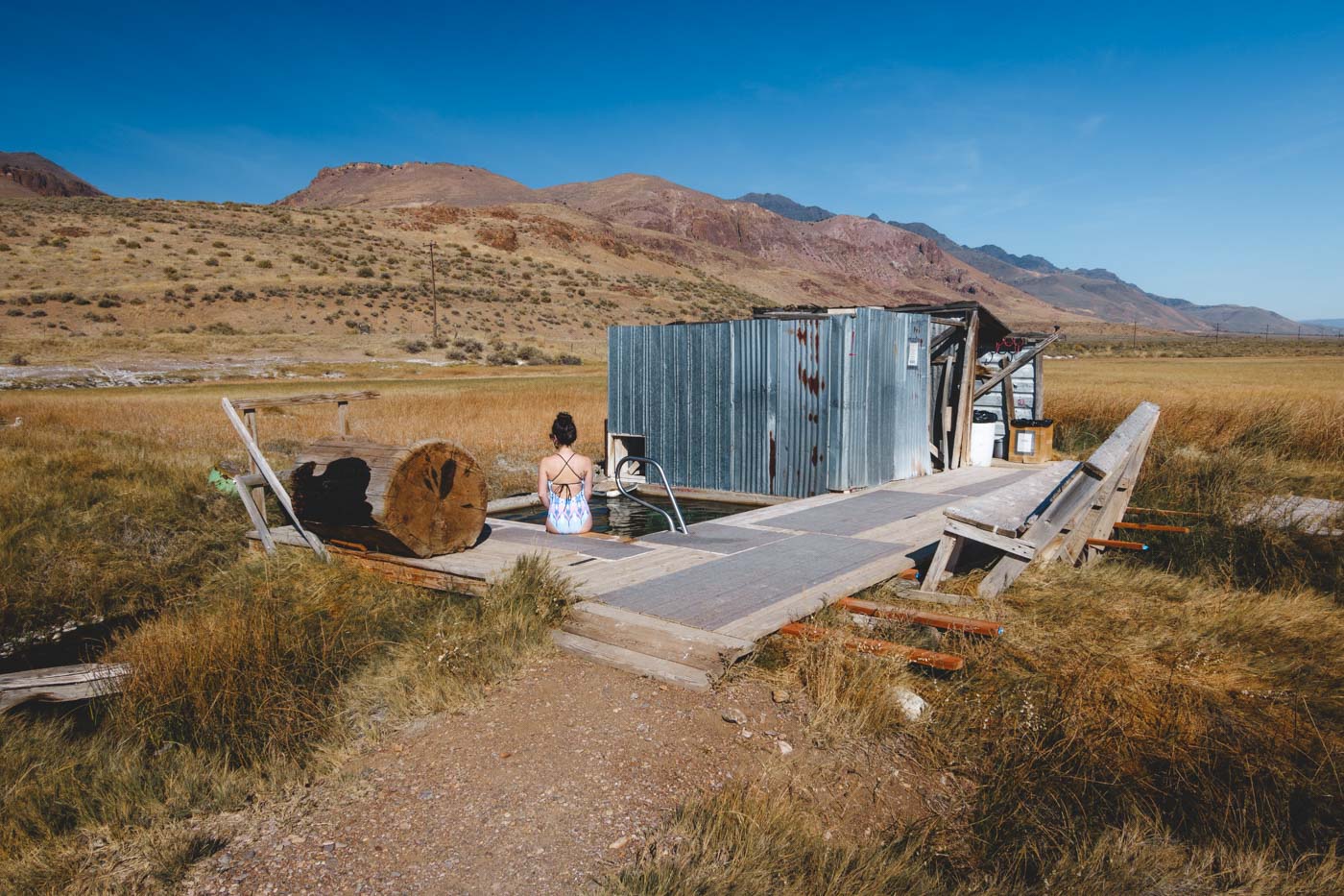 Nina in a bathing suit sitting on the edge of Alvord Hot Springs besides a metal shack with a view of mountains.