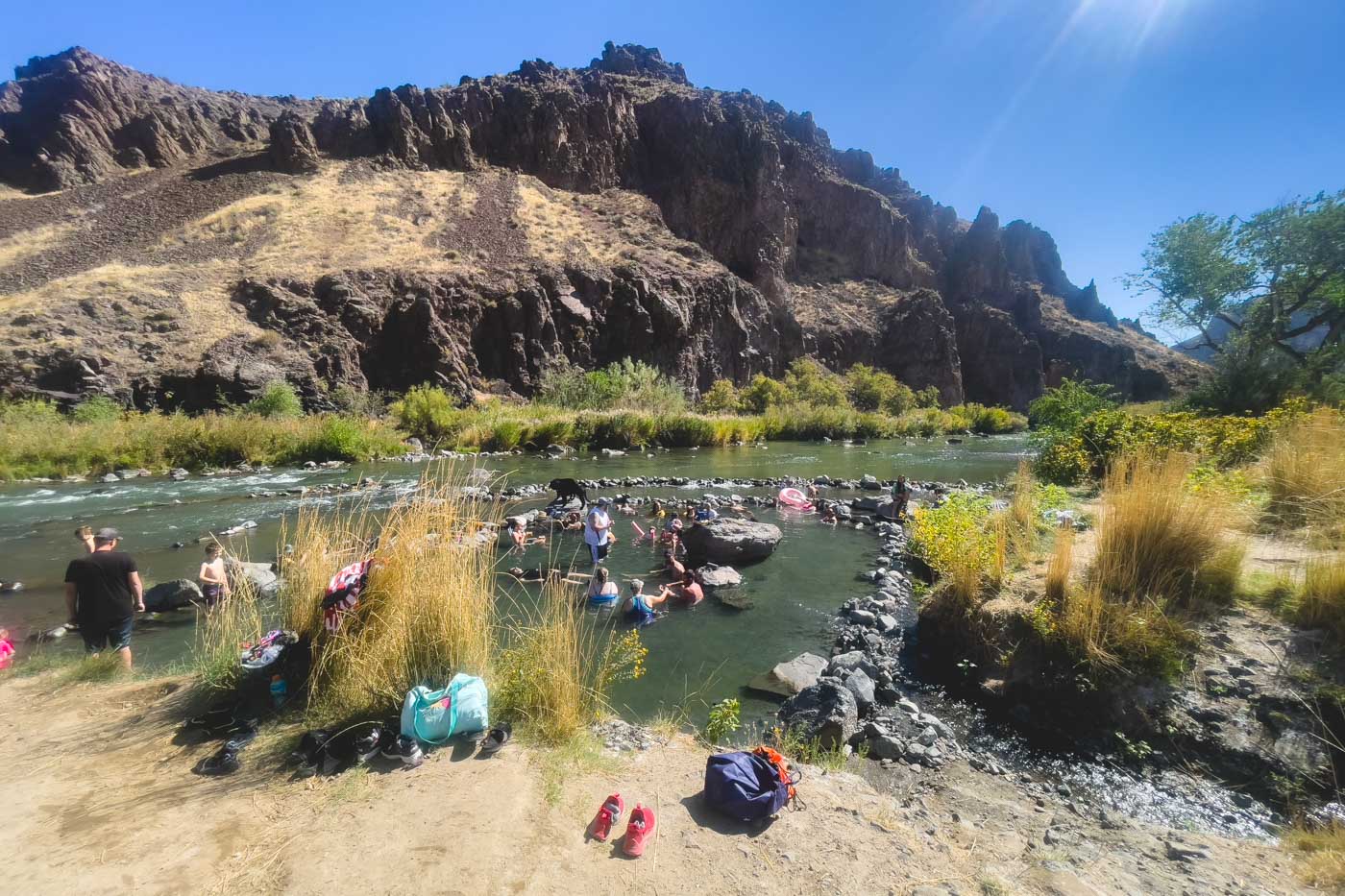 Groups of people bathing in Snively Hot Springs surrounded by bushes, trees and cliffs.
