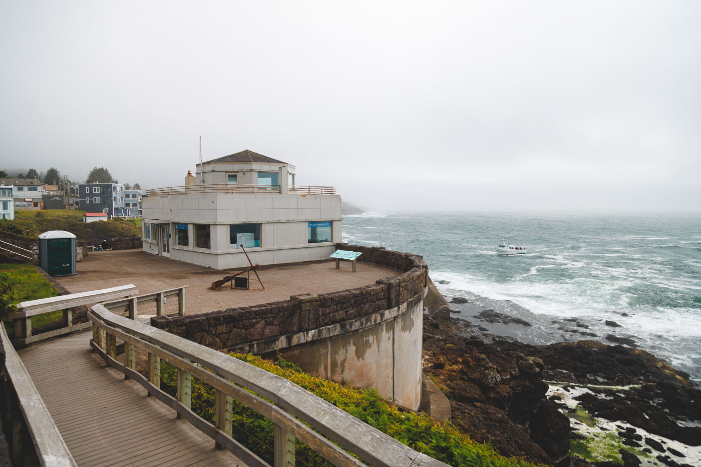 A wooden bridge leading to the white Whale Watching Center overlooking a boat in Depoe Bay on a cloudy day.
