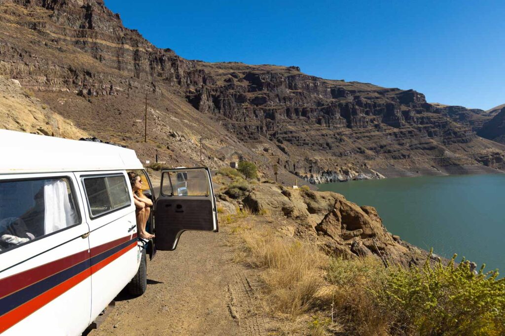 Nina sitting a bit outside her VW Bus that's overlooking Owyhee Lake State Park with dramatic cliffs and the lake below.